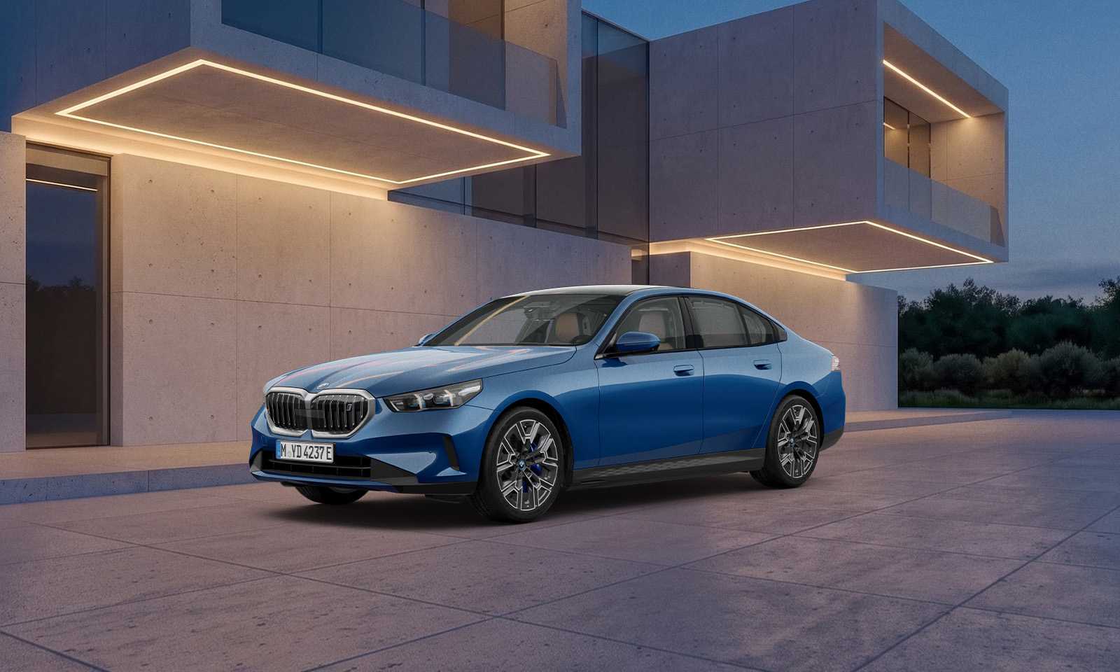 파이토닉 블루 메탈릭 색상의 BMW i5 xDrive40 M 스포츠 에디션이 조명 켜진 콘크리트 건물 앞에 서 있는 앞측면 모습