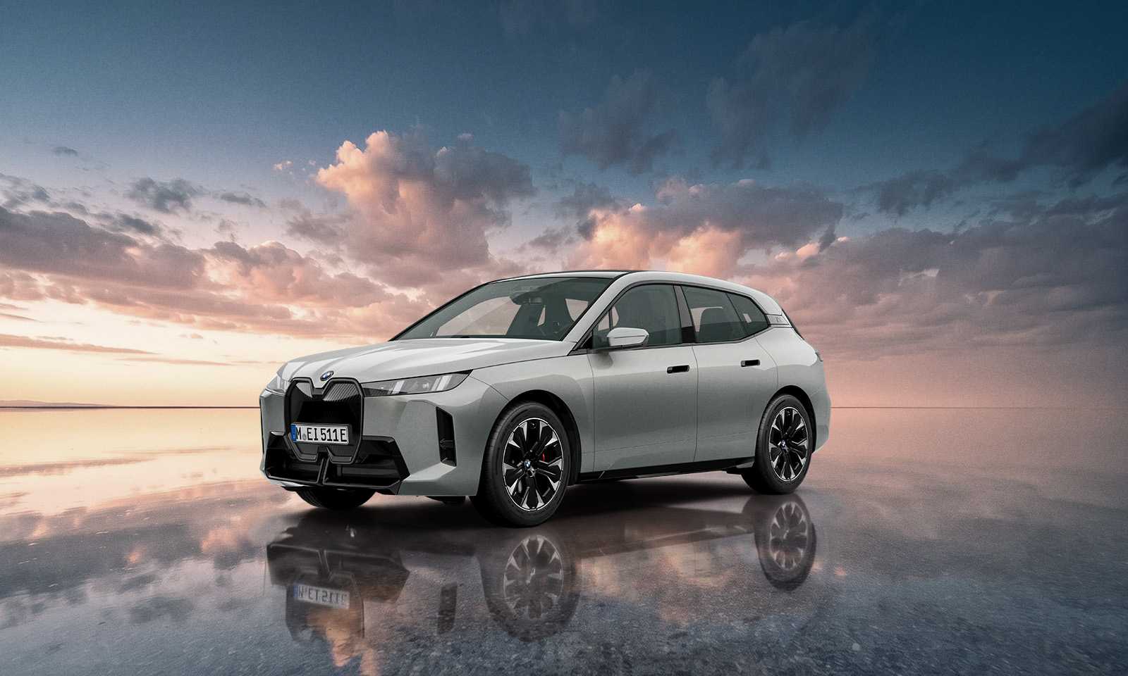 스페이스 실버 메탈릭 색상의 BMW iX xDrive45 M 스포츠 프로 에디션이 노을 진 물가에 비친 앞측면 모습, 빨간 브레이크 캘리퍼